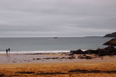 [Overcast day with small cruise ship at sea and two people at water's edge past all the sand.]